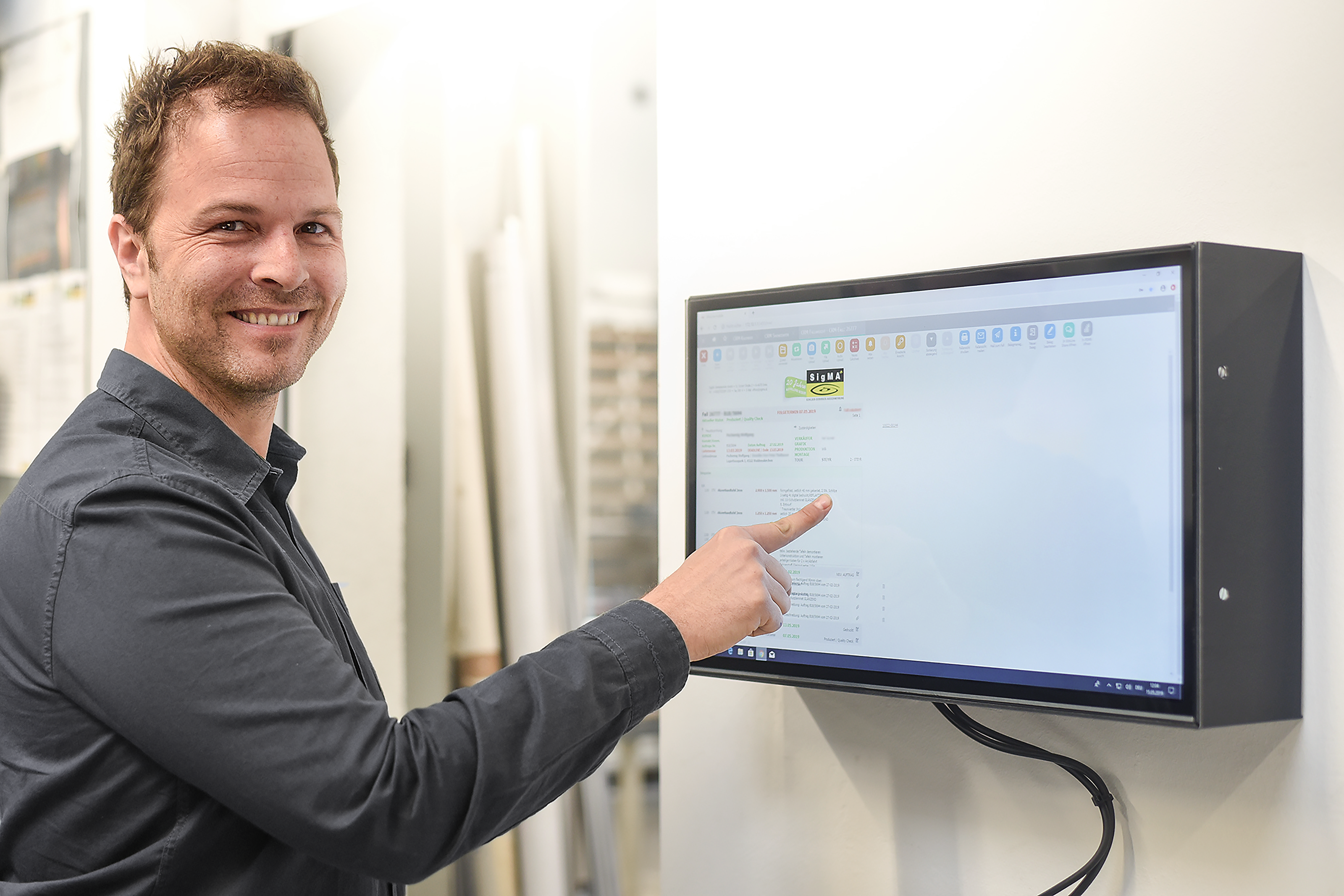 Lächelnder Mann im dunklen Hemd bedient einen an der Wand montierten Touchscreen-Monitor der MIKKA GmbH | © MKKKA GmbH